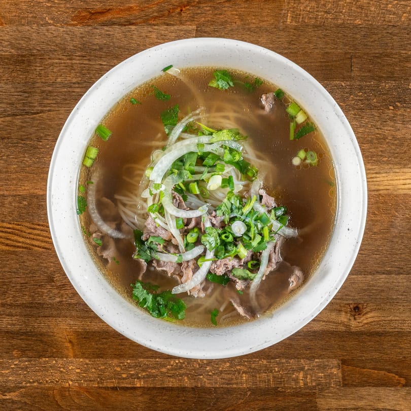 A bowl of Mexiyaki pho with beef, noodles, onions, and herbs in a rich broth.