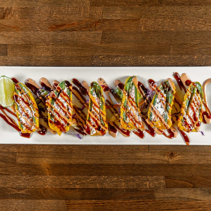 Seven fried jalapeño poppers drizzled with sauces and sesame seeds, served with a lime wedge.