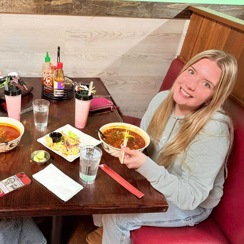 A smiling person enjoying a bowl of Asian soup and dumplings at a table with drinks.
