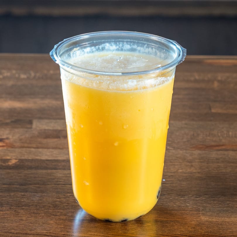 A vibrant yellow mango boba drink in a clear plastic cup, condensation on the outside, on a wooden table.