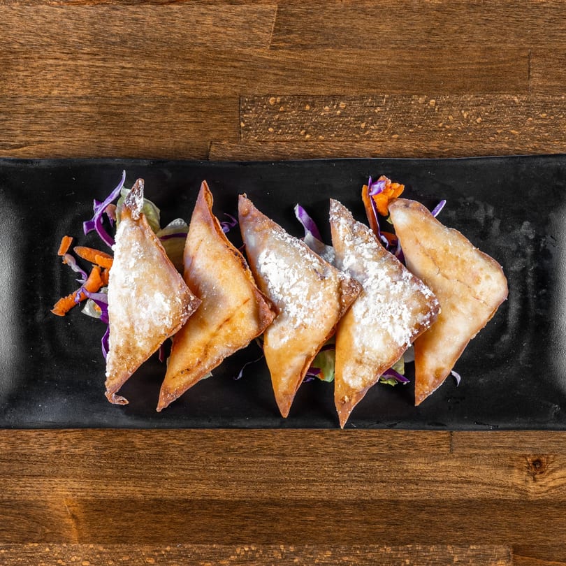 Five golden-brown fried wontons, dusted with powdered sugar, on a black plate with shredded cabbage and carrots.