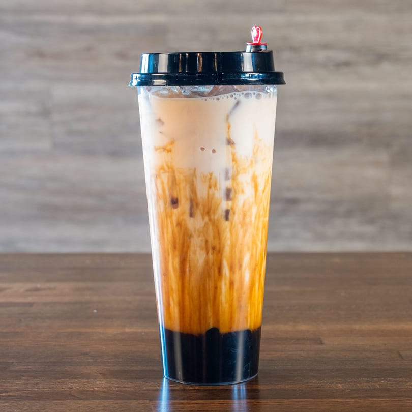 A tall plastic cup of Mexiyaki Asian milk tea with brown sugar swirls and boba on a wooden table.